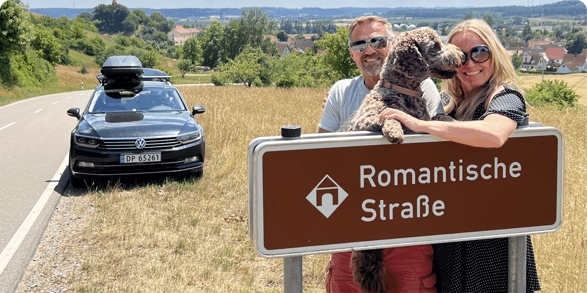 Par med hund ved skilt, bil i bakgrunnen, landskap i Europa. 