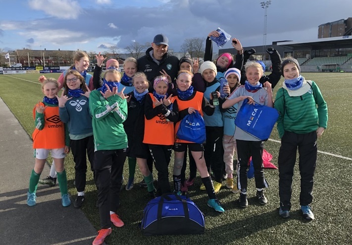 Fotballag feirer på banen med trener, glade barn i sportsutstyr.
