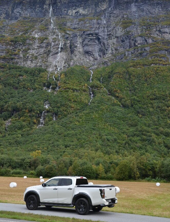 Hvit pickup foran grønn fjellside med fossefall i bakgrunnen.

