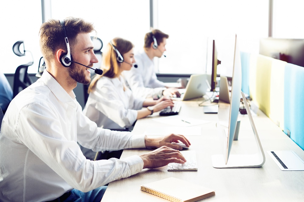 Tre personer med hodetelefoner jobber ved datamaskiner i et lyst kontor.
