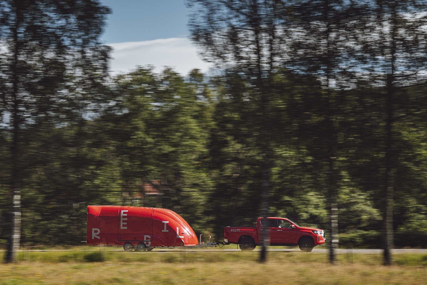 Rød bil og rød skaphenger kjører langs vei med grønne trær