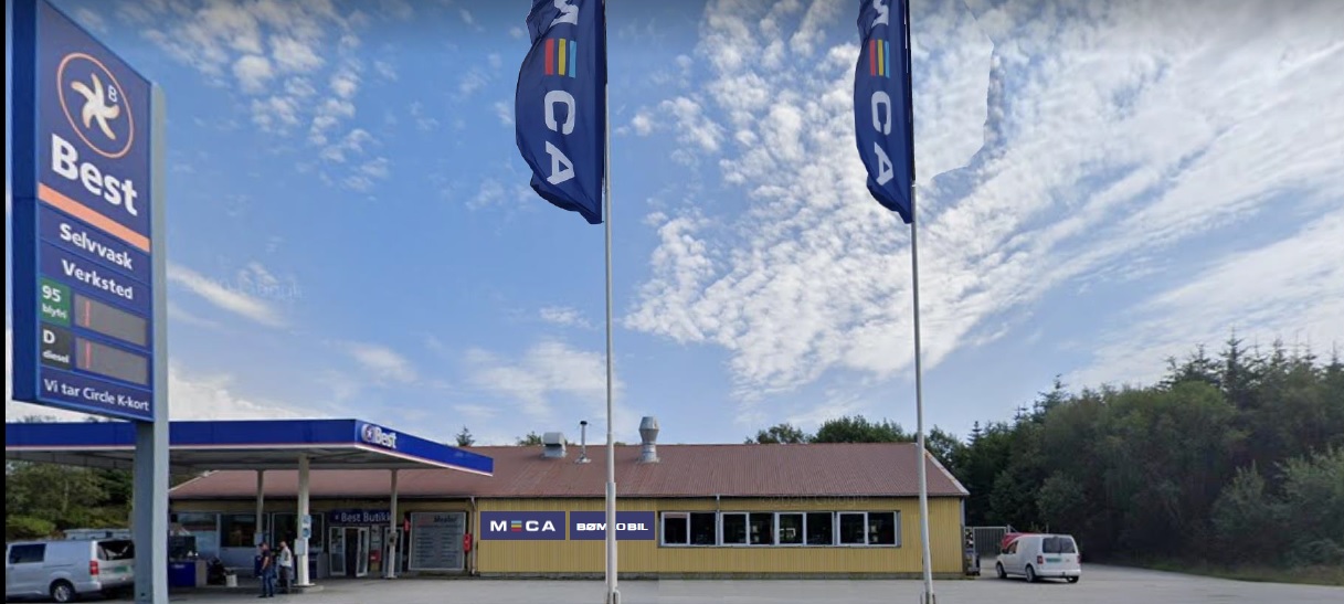 Bilverksted med MECA-flagg, bensinstasjon og parkeringsplass under blå himmel.