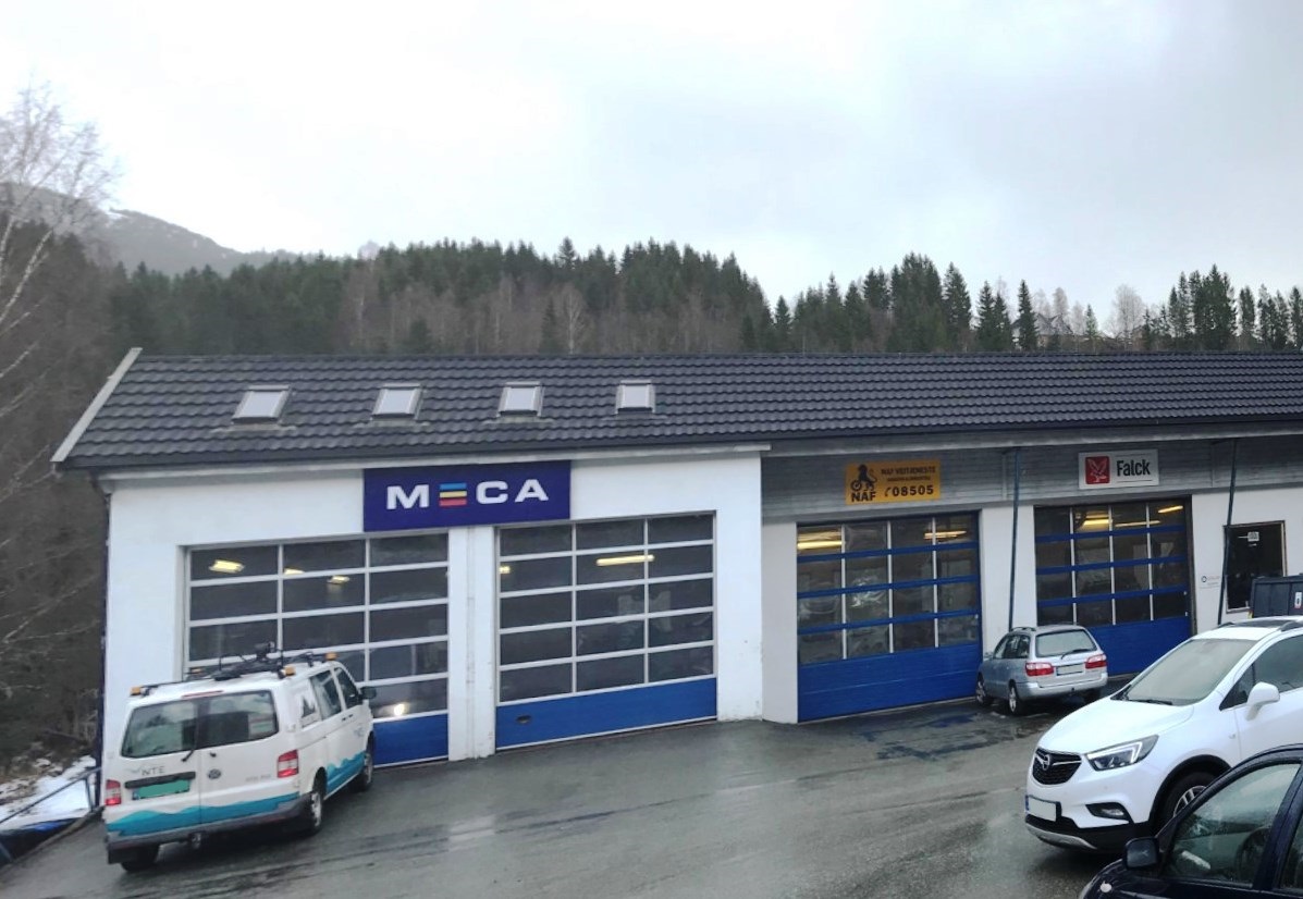 Bilverksted med flere garasjer, omgitt av skog og parkerte biler foran. MECA, NAF og Falck skilt på veggen
