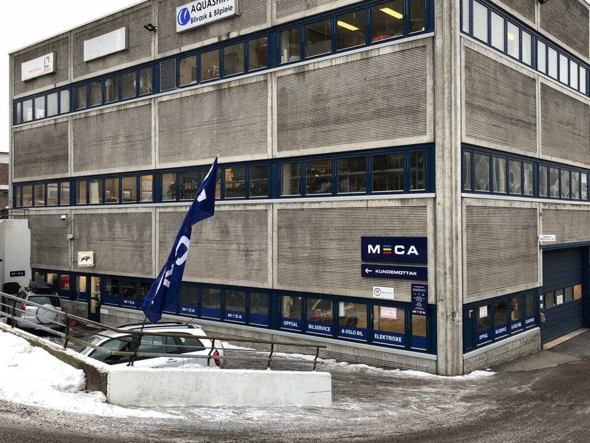 Bilverksted med MECA-skilt, parkert bil, snø på bakken, og flagg utenfor bygningen.