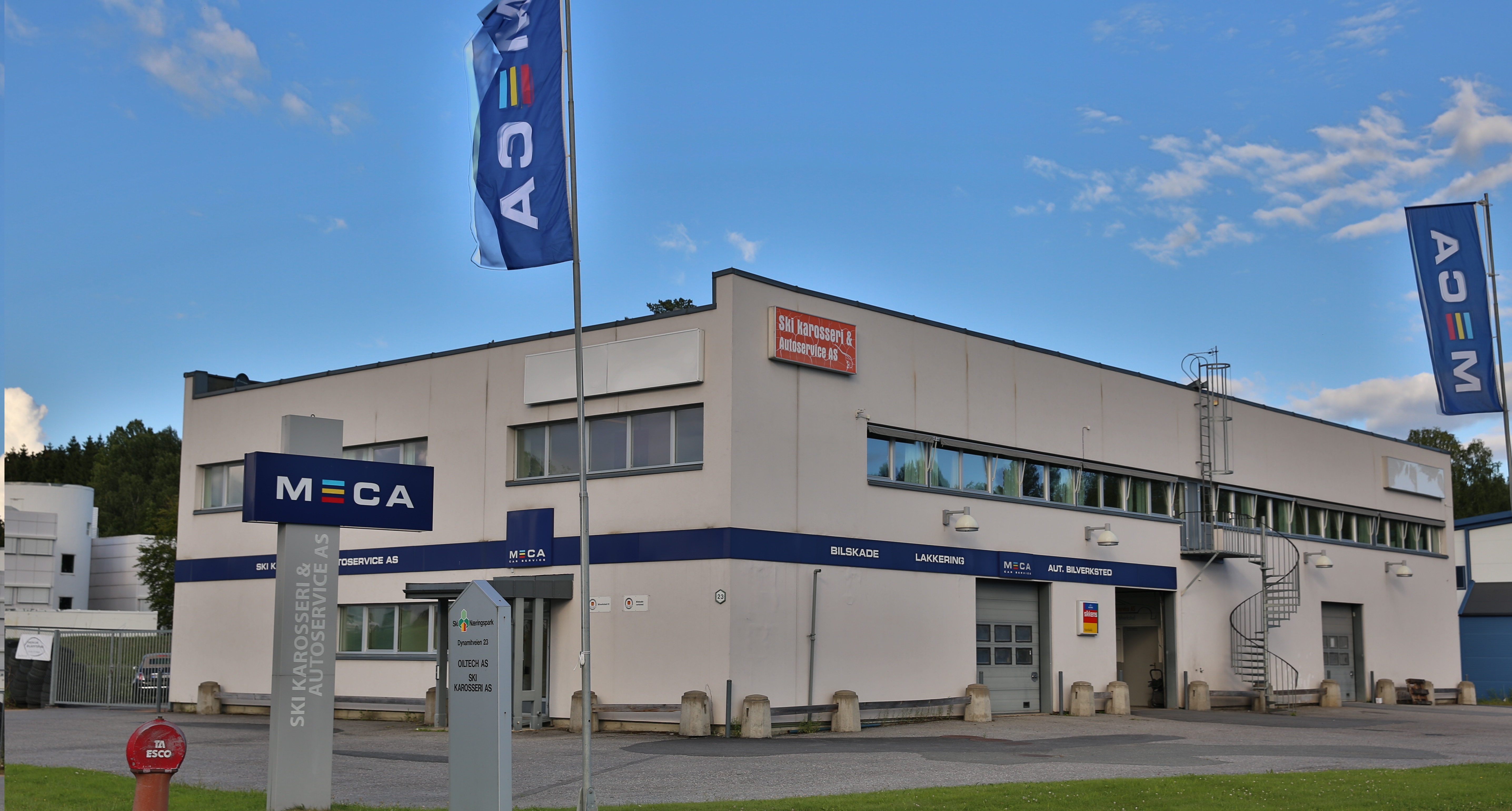 Bilverksted med MECA-skilt, blå himmel, og flagg utenfor bygningen.