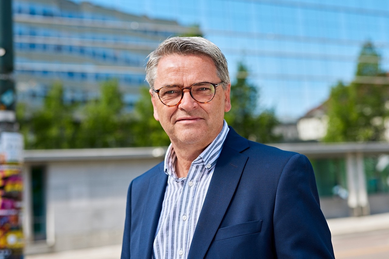 Voksen mann med blå skjorte og dressjakke foran et kontorbygg en sommerdag