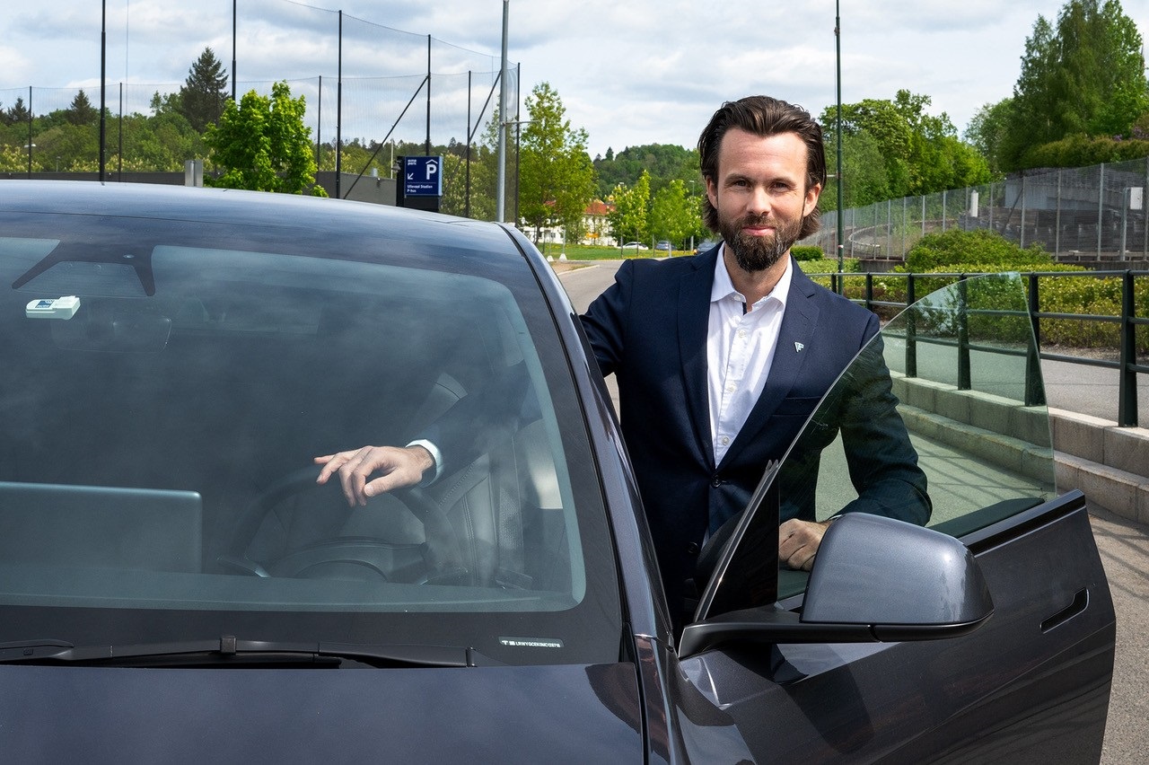 Ung mann i hvit skjorte og svart blazer står i døråpninger til en svart bil. Blå himmel og trær i bakgrunnen
