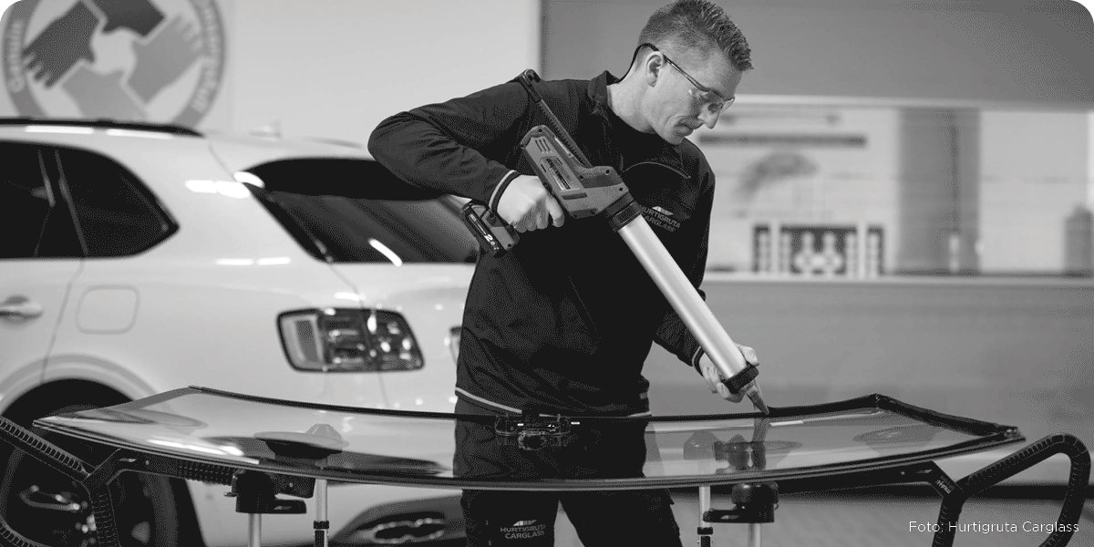 Svart/hvitt bilde av en mekaniker som skifter frontrute på en hvit bil, med teksten Økende bilforsikringspriser -Ny teknologi og større frontruter øker kostnadene