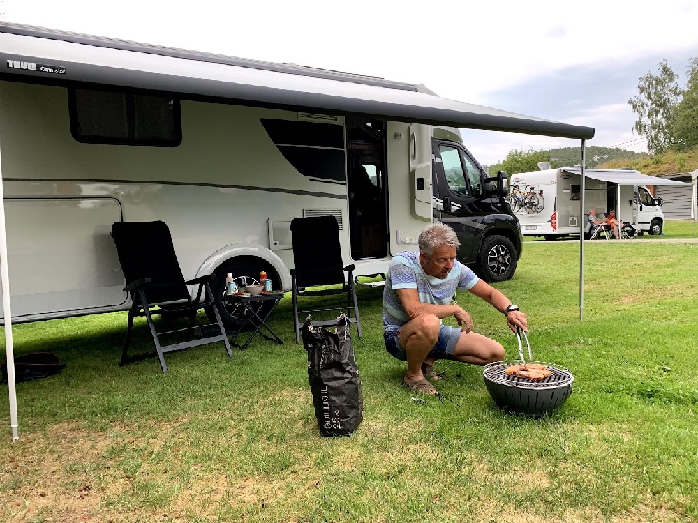 Mann griller foran bobil med campingstoler og bord foran