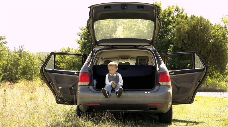 Liten gutt som sitter i bagasjerommet på en parkert bil, omgitt av grønne trør en fin sommerdag