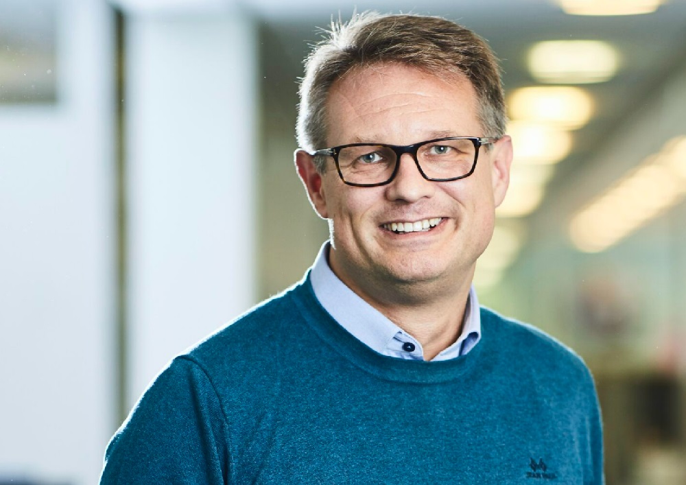 En blid voksen mann med briller og blå genser, med skorte under.