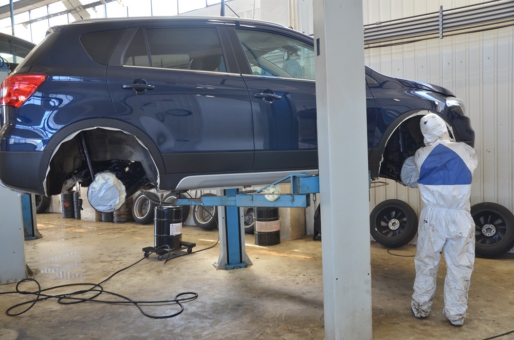 Mekaniker i fullt sikkerhetsutstyr tar antirustbehandling på blå bil, inne på verkstedet