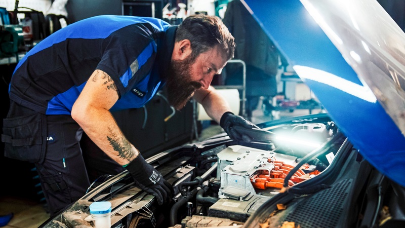Mannlig bilmekaniker med skjegg og blå t-shirt titter ned i motorrommet på en elbil