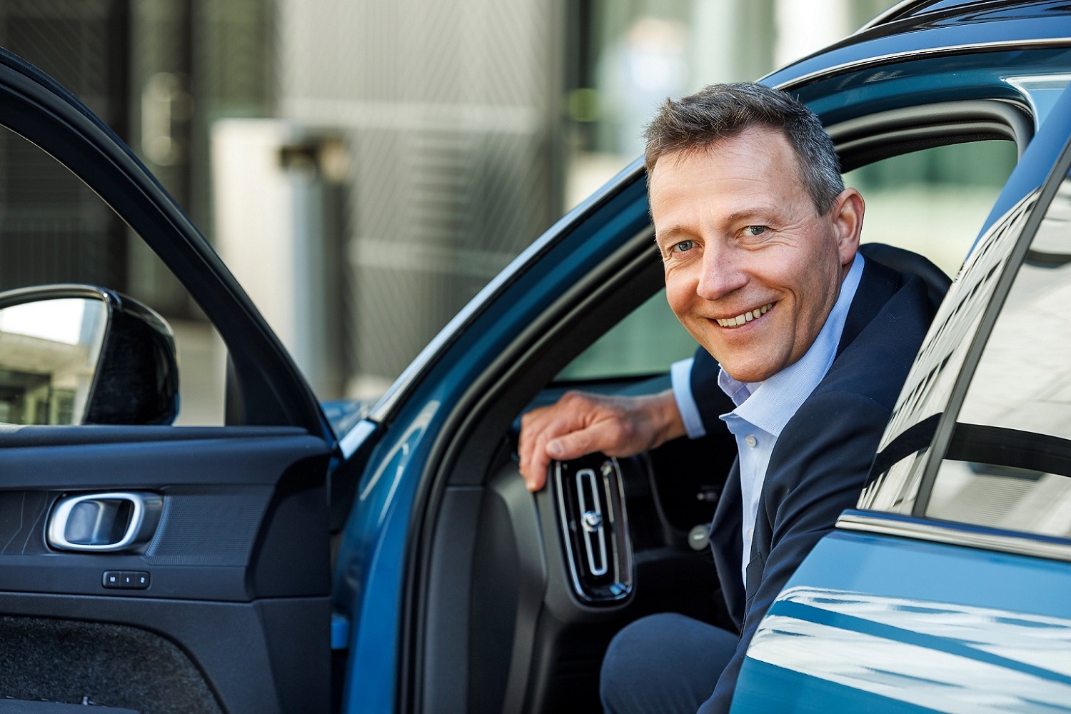 Mann med dress sitter i framsetet på en blå bil med døren åpen