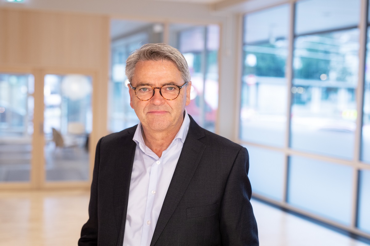 Mann i sort dress og hvit skjorte, med lyst hår og briller i en lyst kontorlandskap med mange vinduer