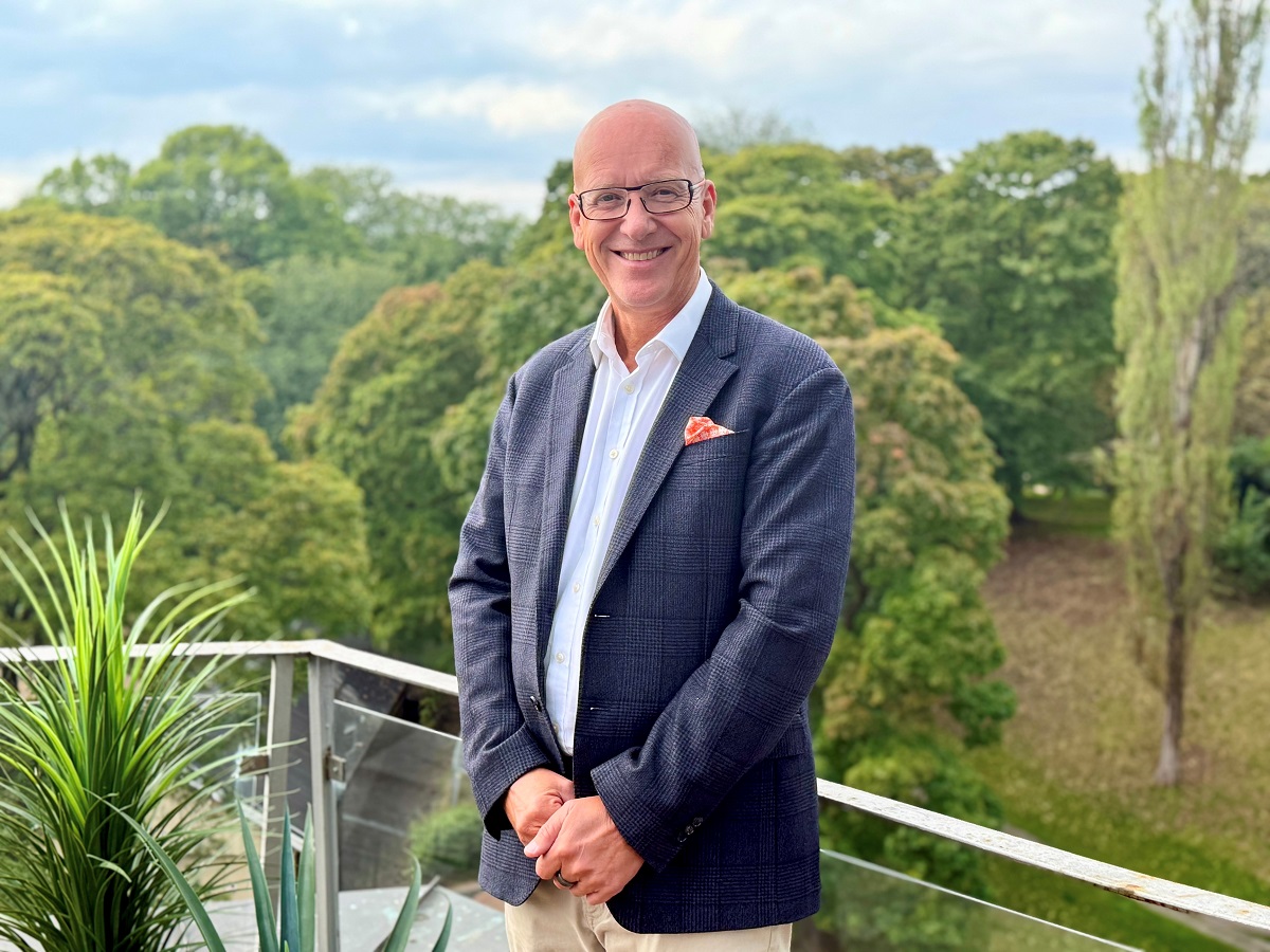 Mann med blå dress står på en balkong med glassrekkverk foran grønne trær