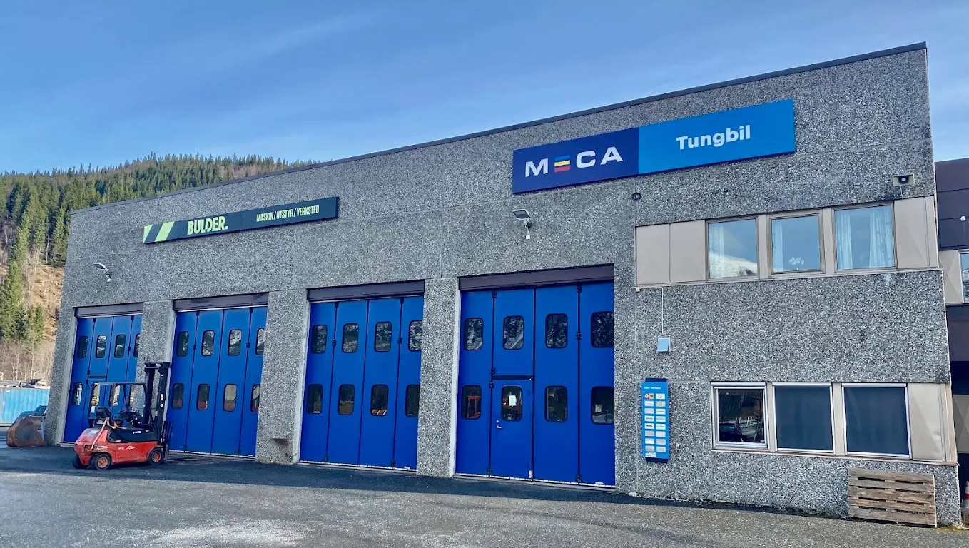 Grått verkstebygg med fire store blå verkstedporter, Med Bulder og MECA Tungbil skilt på veggen