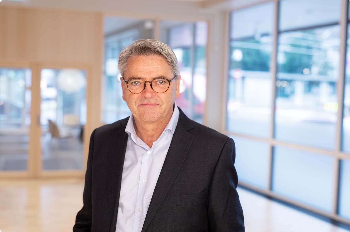 Mann i sort dress og hvit skjorte, med lyst hår og briller i en lyst kontorlandskap med mange vinduer