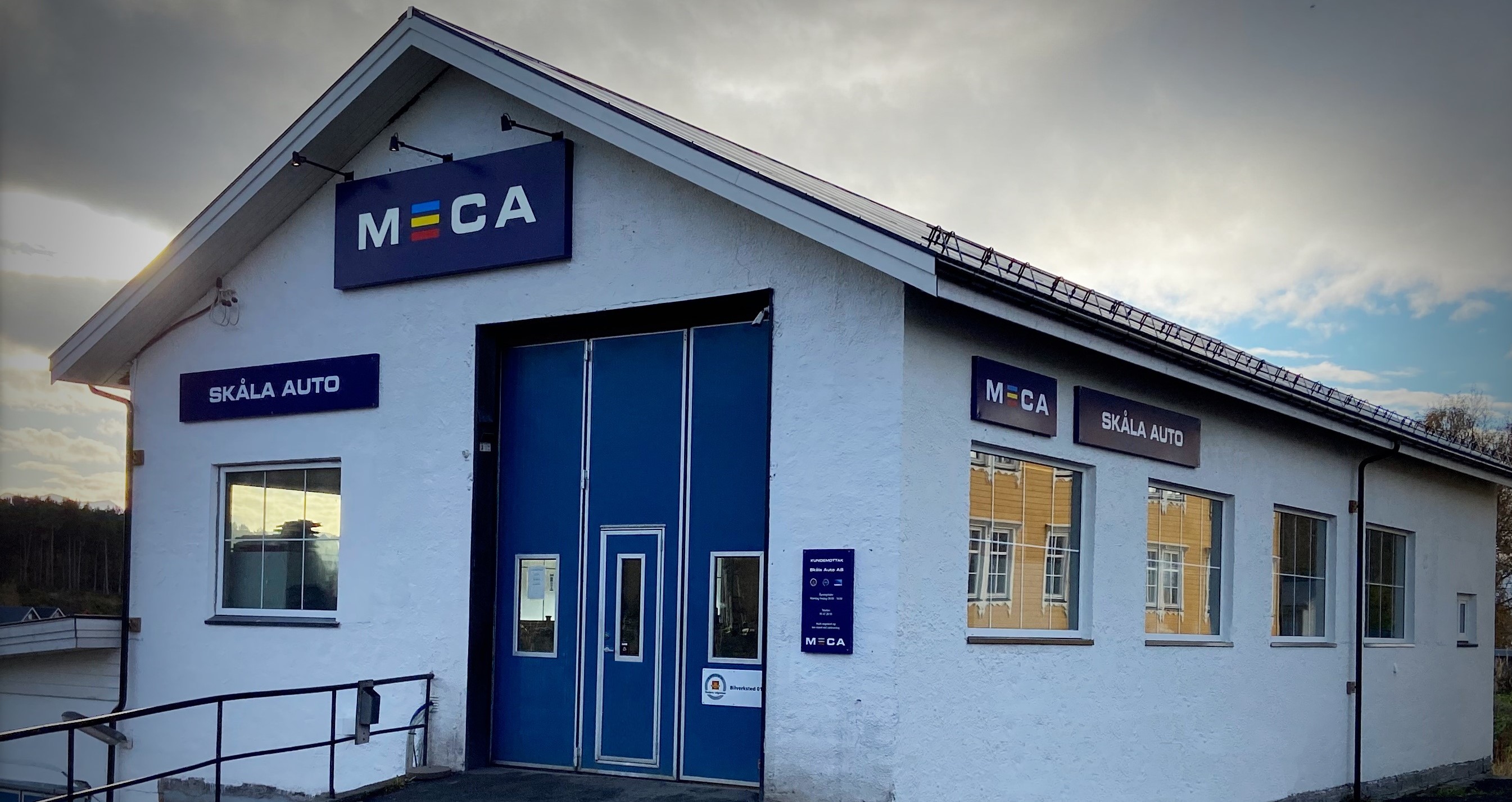 Hvit verkstedbygning i kveldssol, med MECA og Skåla Auto slikt på veggen