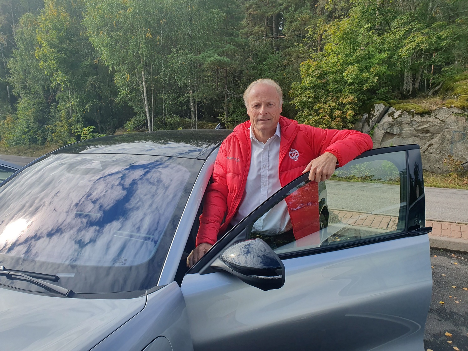 Mann med rød jakke, står i døråpningen på en grå bil.