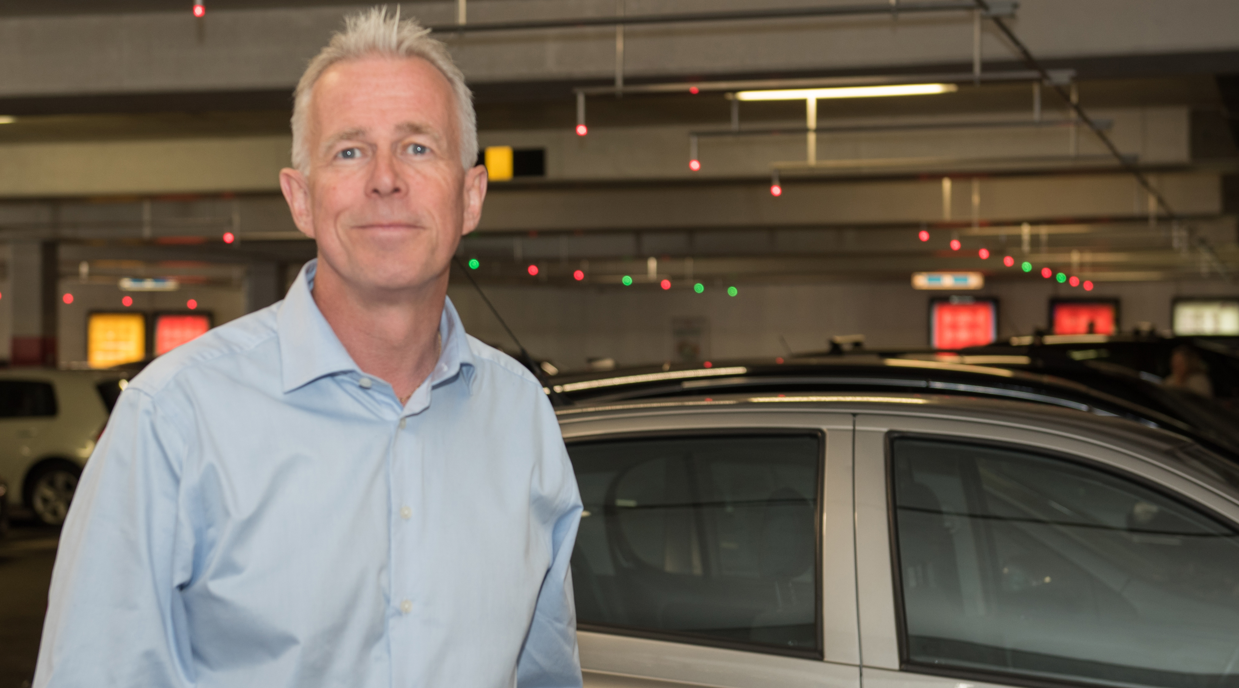 Voksen mann med lysblå skjorte, foran flere biler i et parkeringshus
